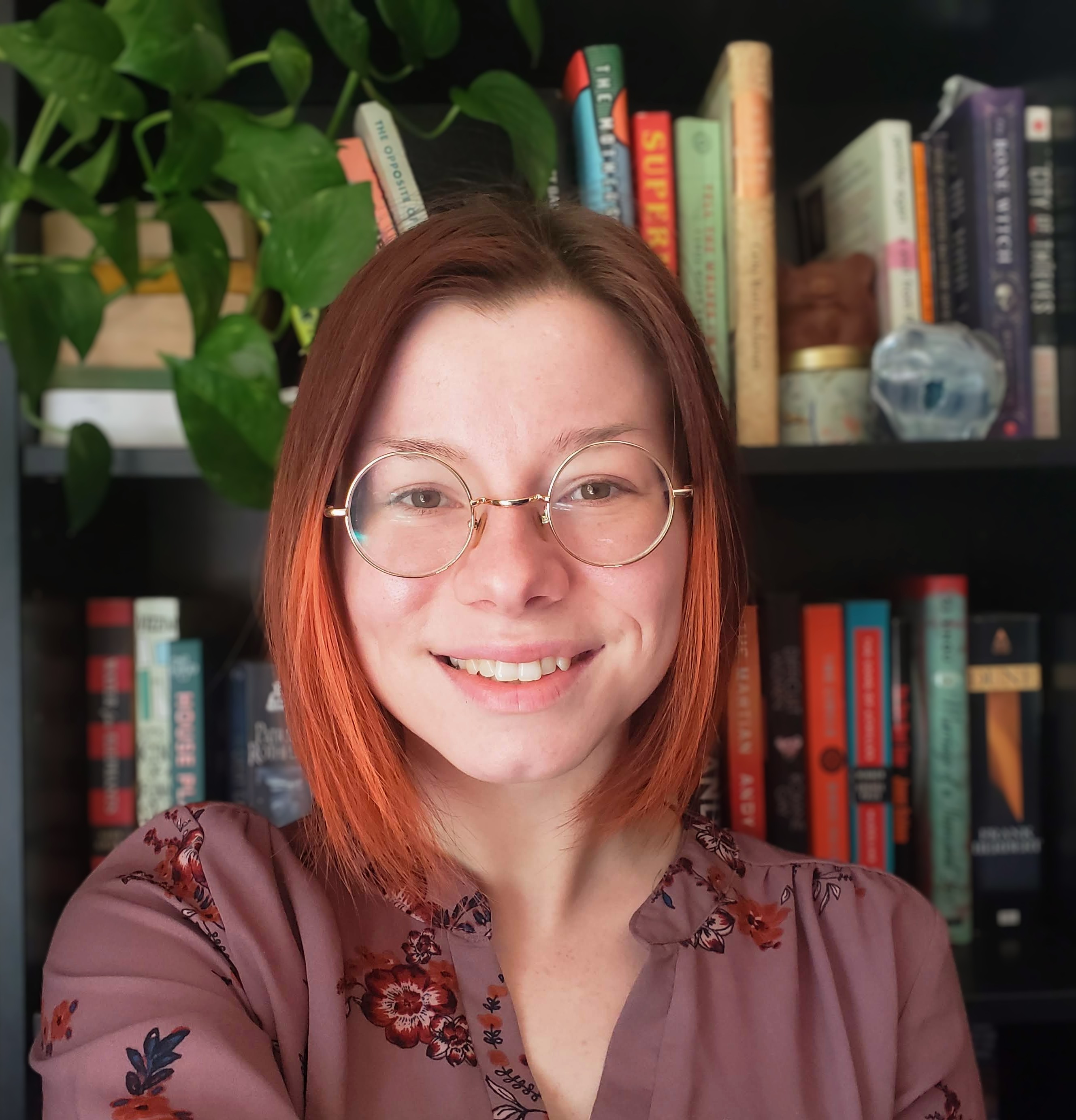 Rhea is a white woman with gold round glasses and a short bob of brown hair with bright orange highlights. She is wearing a mauve colored blouse with a floral pattern.She is posed in front of a full bookcase with a trailing golden pothos on top.  
